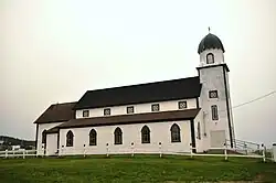 Une église vue de côté. Elle semble de forme basilicale, avec des collatéraux autour de la nef présidée par un clocher au toit arrondi, et une claire-voie. Les murs sont de couleur crème et les atours (toitures, ouvertures et arrêtes) marron