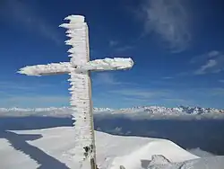 Croix sommitale en bois sur laquelle se sont formés des cristaux de glace dans le sens du vent et une longue chaîne de montagne enneigée en arrière-plan sous un ciel azur.