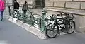 Photographie d'une bouche de métro d'où sortent deux hommes, avec au premier plan une bicyclette ancienne de couleur noire, pourvue d'une étiquette difficilement déchiffrable