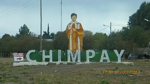 Photo en couleurs d'un panorama semi-rural avec les lettres du village Chimpay surmontées d'une grande représentation du saint