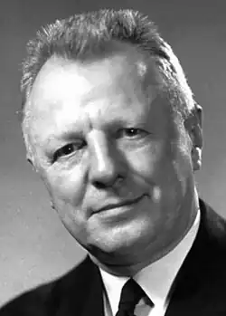 Head and shoulder of a 60-ish man with a flattop haircut and in a coat and tie, looking directly at camera with head tilted to his right and a slight smile.