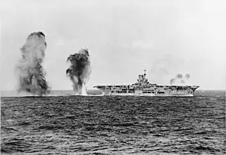 Photo en noir et blanc d'un porte-avions vu de tribord. Des panaches de fumée sont visibles dans l'eau à l'arrière.