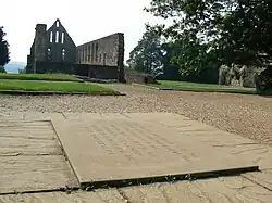 Photo montrant au premier plan une plaque gravée au sol et au fond un édifice religieux en ruine.