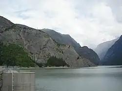Vue depuis un barrage sur un lac où plonge une crête montagneuse, dans une vallée encaissée.