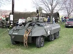 photographie d’un véhicule à roue vu de face avec des camions militaires bâchés à l’arrière plan