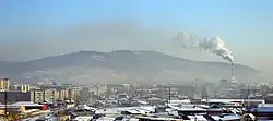 Photographie d'une ville avec des grands ensembles à gauche et des maisons au centre et à droite, le tout recouvert de neige. Au second plan à gauche se distingue une cheminée émettant de la fumée. Un smog épais stagne au-dessus de la ville, qui cache partiellement la montagne en arrière-plan.
