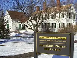 Photographie d'une maison blanche à deux étages avec de hautes cheminées en brique. Le sol est recouvert de neige et un panneau indique qu'il s'agit de la résidence du 14e&nbsp;président des États-Unis.