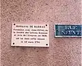Plaque de couleur blanche à écriture noire rendant hommage à Hippolyte de Barrau pour la fondation de la Société des lettres, sciences et arts de l'Aveyron. À sa droite, une plaque bleue indiquant la « rue Neuve ».
