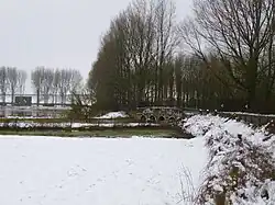 Photo d'un paysage enneigé. Au deuxième plan, on voit les arches d'un pont qui enjambe une rivière. À l'arrière se dressent des arbres nus.