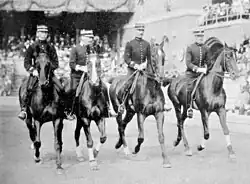 Photo noir et blanc. Devant une tribune, quatre cavaliers avancent alignés de front, vus de face. Tous sont des militaires.