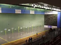 Vue de l'intérieur d'une salle de sport, à gauche un terrain de pelote basque, à droite des tribunes.