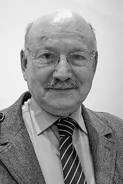 Un homme en costume, moustache, crâne dégarni et lunettes rondes, regarde devant lui.