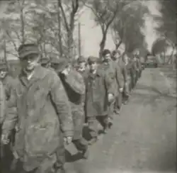 Photo en noir et blanc montrant une file d'hommes en uniforme marchant sur une route entourée d'arbres. Dans le fond, une jeep est visible. Un homme au premier plan regarde l'appareil photo.