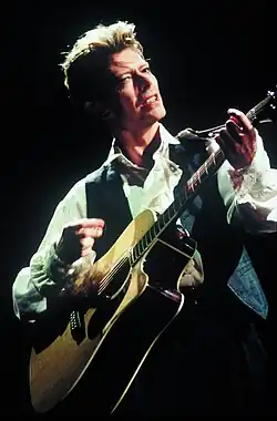 Photo d'un homme blanc aux cheveux blonds coupés courts. Il porte une chemise blanche et une veste sans manches noires et joue de la guitare acoustique.