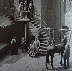 Photo en noir et blanc d'une cour d'immeuble, un cheval avec son propriétaire se trouvant au bas à droite de la photo, une femme et des enfants se tenant dans la cour, et trois hommes se tenant en haut des marches d'escalier.