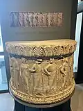 Socle semi-circulaire en marbre, probablement un élément décoratif de l'autel d'Apollon au Monument d'Auguste. On y voit représentés divers dieux et héros du panthéon grec ; on notera le style archaïsant.