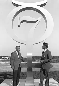 Photographie en noir et blanc de Grissom et Schirra à côté d'un monument représentant l'emblème du programme Mercury.