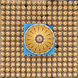 au centre un carré bleu, écorné à un angle, avec des rayons en forme de flèche qui partent du centre. Tout autour, bien alignés, de nombreux petits cercles jaunes, avec une sorte de pic dessus.