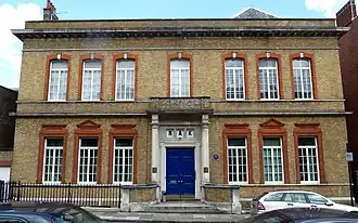 Facade d'un bâtiment symétrique de couleur grise, orné de 13 fenêtres avec une grande porte bleue au milieu.