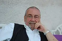 Portrait buste d'un homme assis grisonnant portant des lunettes, chemise et gilet dans une pause décontractée.