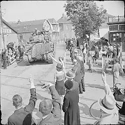 photographie en noir et blanc d’un char traversant un village et acclamé par une foule