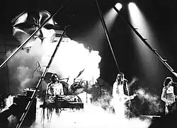 Photo en noir et blanc de trois musiciens sur une scène envahie de fumée blanche. Celui de gauche est derrière un synthétiseur, les deux autres ont des guitares. À l'arrière-plan apparaît un immense visage d'apparence égyptienne.