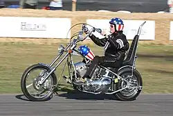 Photo d'un homme assis sur une moto au guidon démesurément grand ; son casque et le corps de la moto sont décorés aux couleurs du drapeau des États-Unis