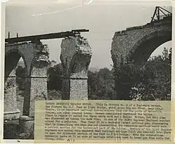 Vue en plongée du pont dynamité, deux arches sont manquantes, le tablier temporaire est en cours d'installation, reposant sur uniquement deux piles du pont.