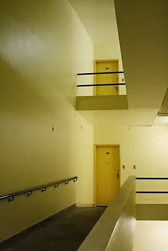 Vue de l'intérieur. On voit deux portes sur deux étages dans un couloir jaunâtre.