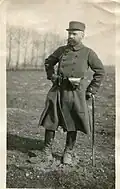 Photo noir et blanc d’un homme en uniforme, les chaussures pleines de boue dans un paysage de campagne, qui pose appuyé sur une canne.