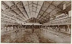 Vue du grand bassin de la piscine avec des nageurs sous un toit du type halle. Au fond du bassin sur le petit côté se trouvent des gradins.