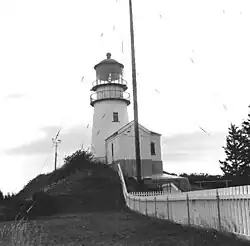 Photo en noir et blanc du phare vu de près, on ne voit pas les environs. La peinture blanche du phare est propre, un grand poteau est planté à l'entrée et supporté par des haubans.