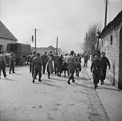 Photo noir et blanc montrant des hommes en uniforme et armés qui encadrent des civils dans une rue.