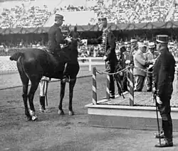 Photo noir et blanc. Le roi, en grand uniforme, de profil sur une estrade, tend la médaille à Cariou, qui est à cheval, vu de dos.