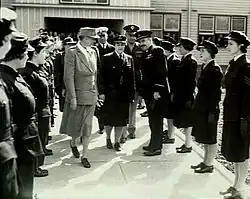 Photographie en noir et blanc représentant deux femmes et trois hommes entourés de militaires vêtus de costume sombre.