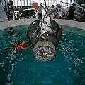 Photographie en couleur de Stafford et Shirra dans une piscine avec une reproduction du vaisseau Gemini.