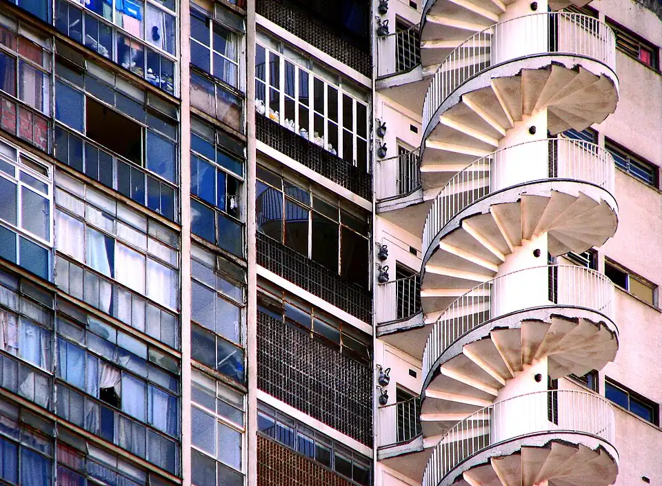 Gros plan sur la façade en béton et en verre du bâtiment, permettant de voir l'escalier blanc en colimaçon qui court le long de l'édifice.