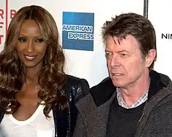 Photo en gros plan d'une femme noire aux longs cheveux portant un haut blanc et une veste noire et d'un homme blanc aux cheveux courts portant une chemise à carreaux, un gilet et une veste.
