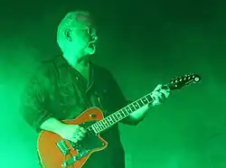 Photo d'un homme blanc à lunettes, avec des cheveux blancs coupés courts et une barbiche également blanche. Il porte une chemise noire et joue d'une guitare électrique rouge.