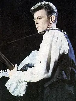 Photo d'un homme blond portant une chemise blanche à froufrous et un veston noir. L'air concentré, il joue d'une guitare acoustique suspendue autour de son cou.