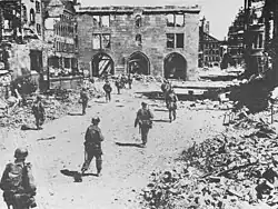 Photo noir et blanc montrant des soldats, de dos, marchant dans une rue entourée de bâtiments en ruine et éventrés.