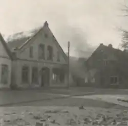 Photographie en noir et blanc montrant un bâtiment avec de la fumée et des flammes s'échappant du toit et de l'intérieur. Sur la façade est écrit "Friseur", signifiant "coiffeur" en allemand.
