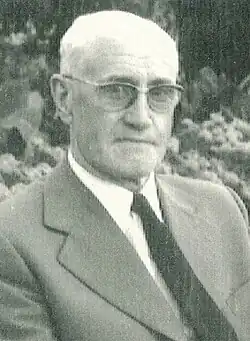 Photographie en noir et blanc d'un homme âgé porteur de lunettes, qui regarde l'objectif sans sourire.