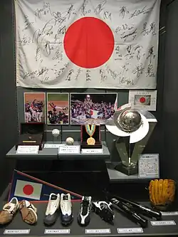 Photo en couleur d'une vitrine, au premier plan des chaussures, gants et battes de baseball, au deuxième rang des médailles et un trophée, à l'arrière plan un drapeau blanc et rouge