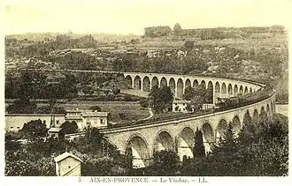 Photographie en sépia montrant un train composé d'une locomotive coupe-vent de de plusieurs voitures sur le pont, il décrit une trajectoire en courbe et la campagne autour du viaduc est peu urbanisée.