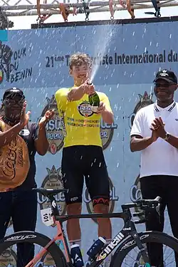 Jeune homme blanc vêtu d’un maillot jaune et d’un cuissard noir tenant une bouteille de proseco ouverte, accompagné de deux hommes noirs avec des lunettes de soleil et des casquettes noires.