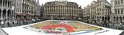 Vue panoramique d'une planche géante montrant une fusée sur sa tour de montage, au centre de la Grand Place de Bruxelles, les spectateurs étant séparés de l'image par des barrières.