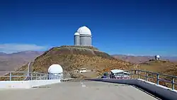 Le site de l'observatoire de La Silla vu depuis l'emplacement du NTT (New Technology Telescope). Au centre, la grande coupole abrite le télescope de 3,6 mètres de l’ESO, accompagné sur sa droite de l’ancienne coupole du télescope auxiliaire Coudé (CAT), reliée par une passerelle. À droite de l'image, sur une colline secondaire, est visible l’antenne argentée du radiotélescope SEST (Swedish-ESO Submillimetre Telescope), qui a été mis hors service en 2003. Le SEST a été remplacé par des installations plus modernes telles que l'APEX et l'ALMA, situées sur le plateau de Chajnantor (Chili).