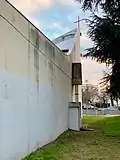 Vue de côté d'un bâtiment en béton, avec des traces d'humidité ; le bâtiment se prolonge avec une partie montante surplombée d'une croix.
