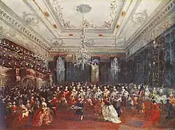 Haute salle éclairée par trois lustres, pleine de gens debout ; sur un côté se trouvent des gradins avec les musiciens.